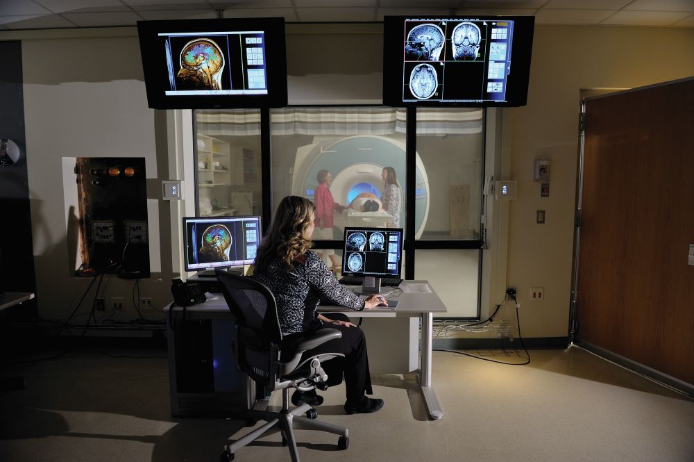 An MRI tech looks at scans while another works with a research subject in the background